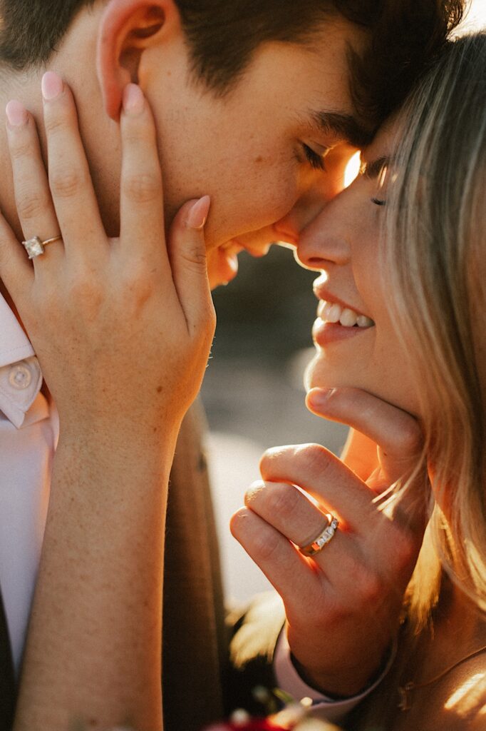 A bride and groom smile as they are about to kiss during sunset, each of their hands rest on the other one's face showing off their wedding rings