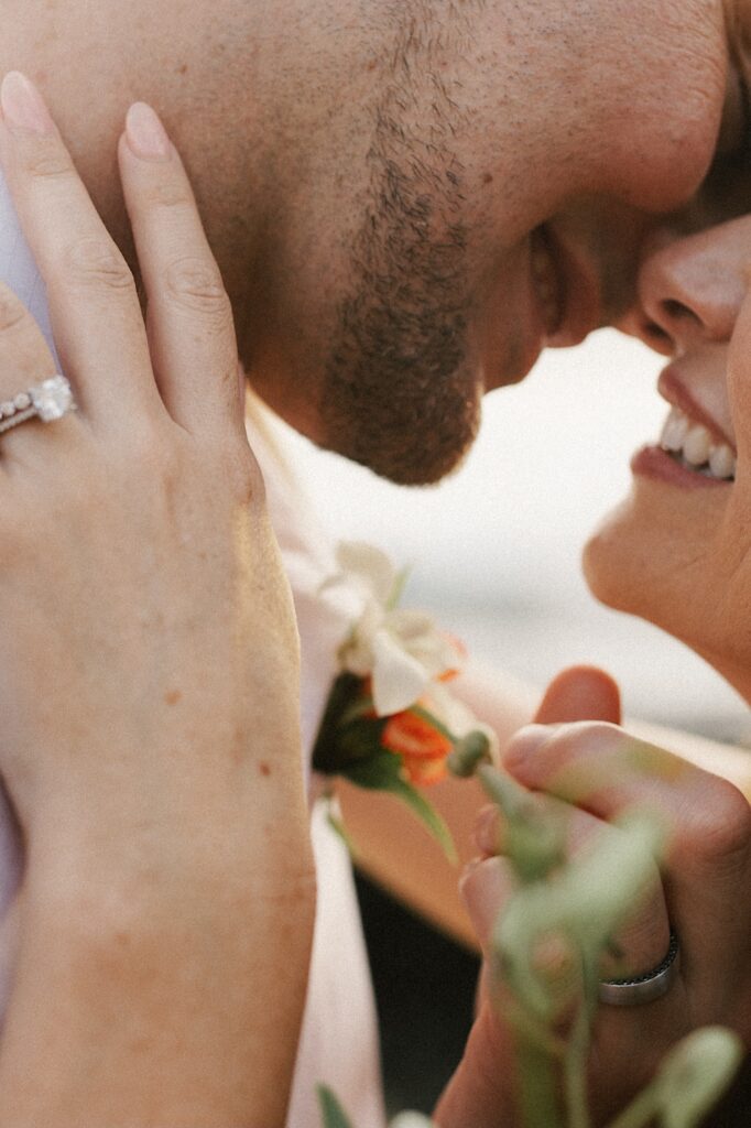Close up photo of a man and woman smiling while about to kiss, their hands are also in the photo each with their wedding ring on it