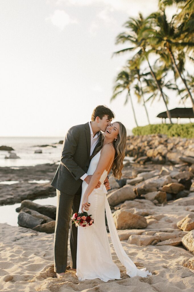 A bride smiles and laughs as the groom kisses her on the cheek while they stand together on a beach at sunset with palm trees and the ocean behind them