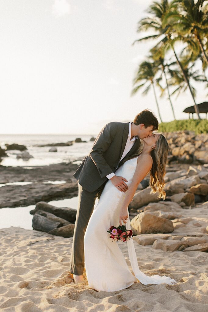 A bride and groom kiss one another while standing on a beach at sunset with palm trees and the ocean behind them