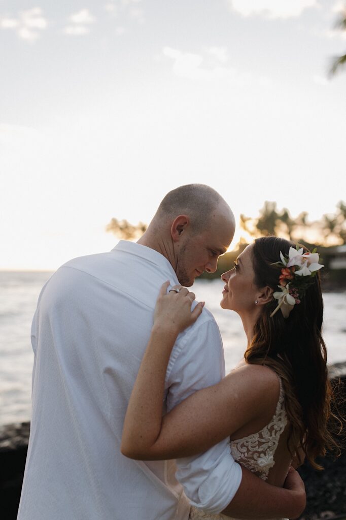 A bride and groom stand side by side and look at one another as the sun sets on the ocean behind them