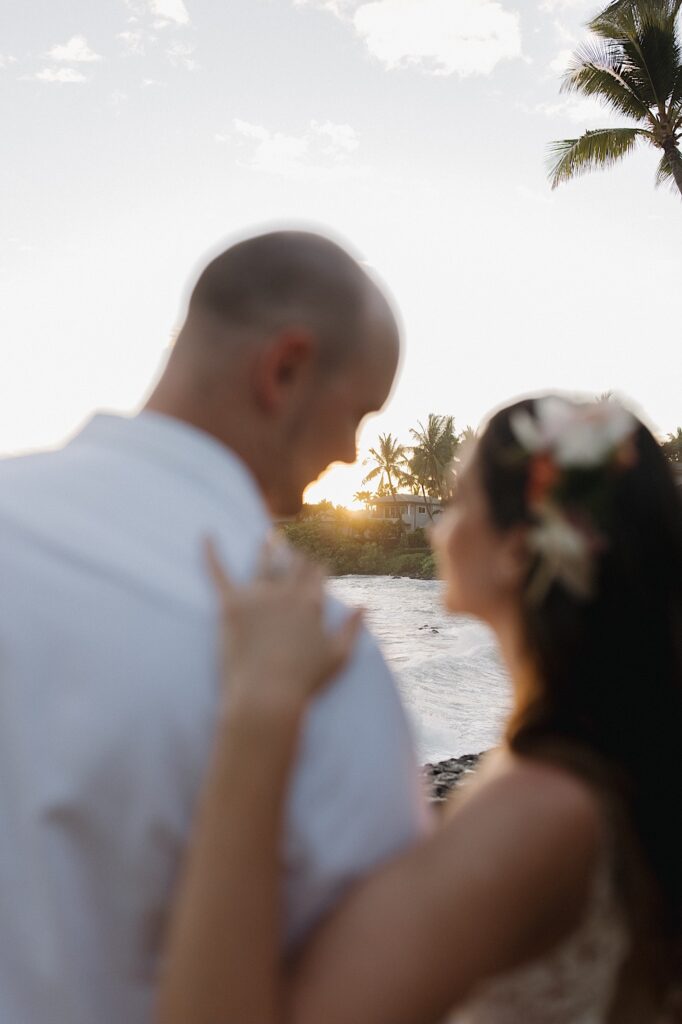 A bride and groom stand next to one another and look out at the sunset in the background