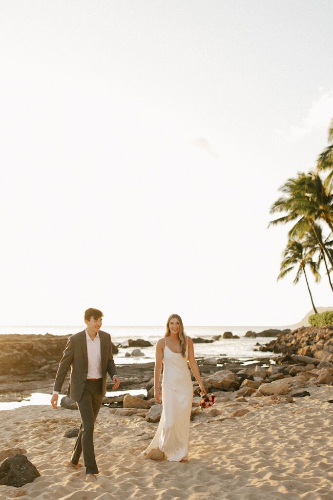 A bride and groom walk next to one another on a beach and smile at the camera with the ocean and palm trees behind them