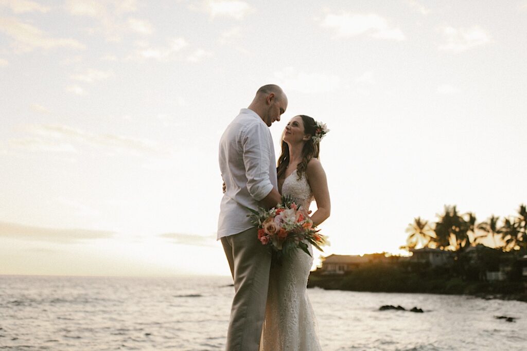 A bride and groom stand facing and holding one another while smiling at each other after their intimate wedding ceremony on Hawaii's big island, behind them is the ocean during sunset
