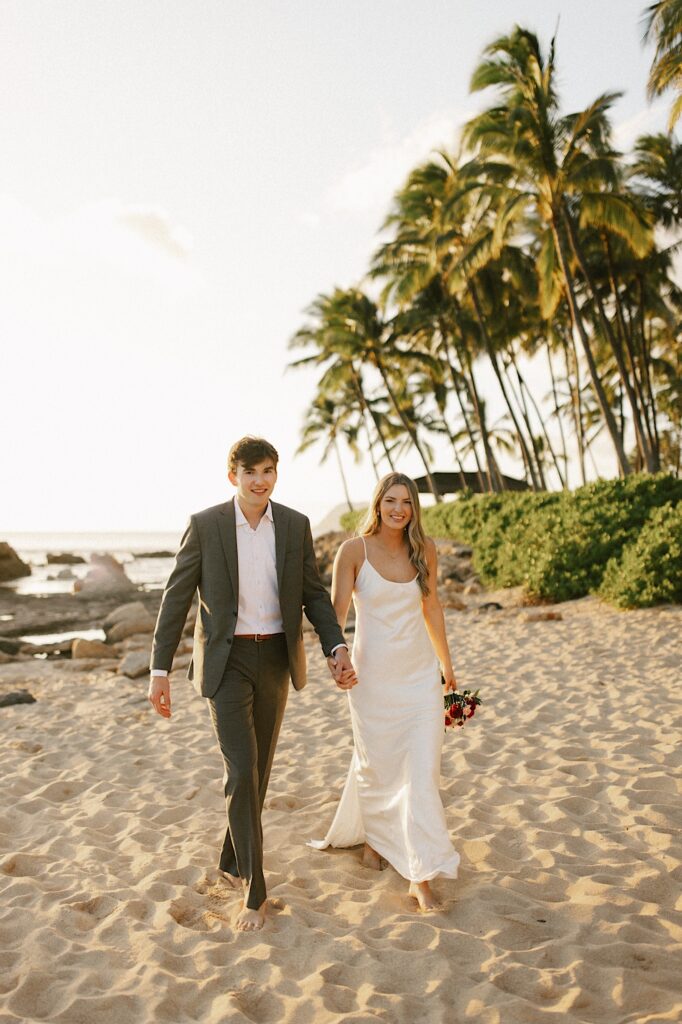 A bride and groom hold hands and walk on a breach towards the camera while smiling during sunset with the ocean and palm trees behind them