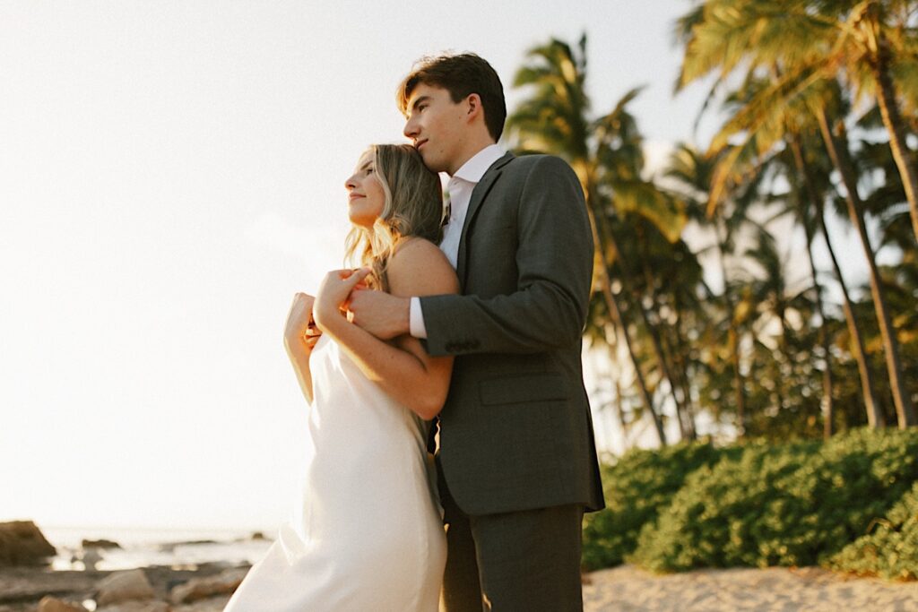 A groom stands behind a bride and holds her while the two look out at the ocean together while the sun sets during their elopement at Lanikūhonua on Oahu, they're standing on a beach with palm trees in the background