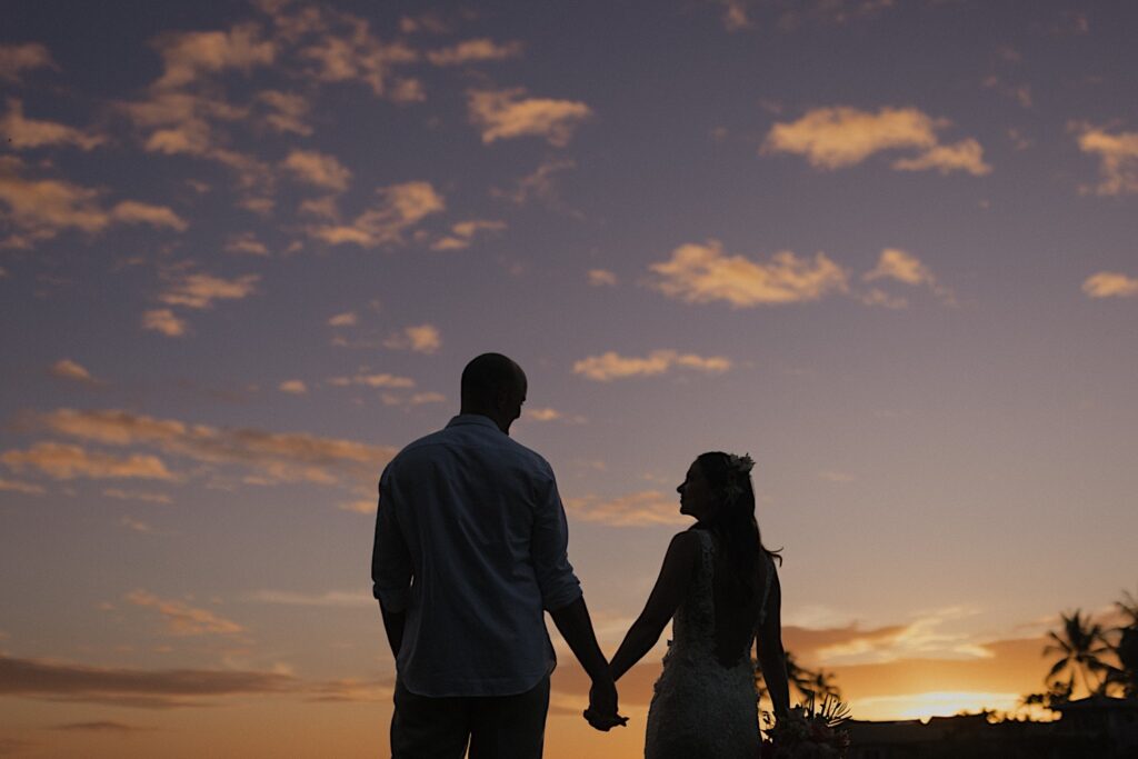 Silhouette of a bride and groom standing next to one another and holding hands while looking at each other as the sun sets behind them