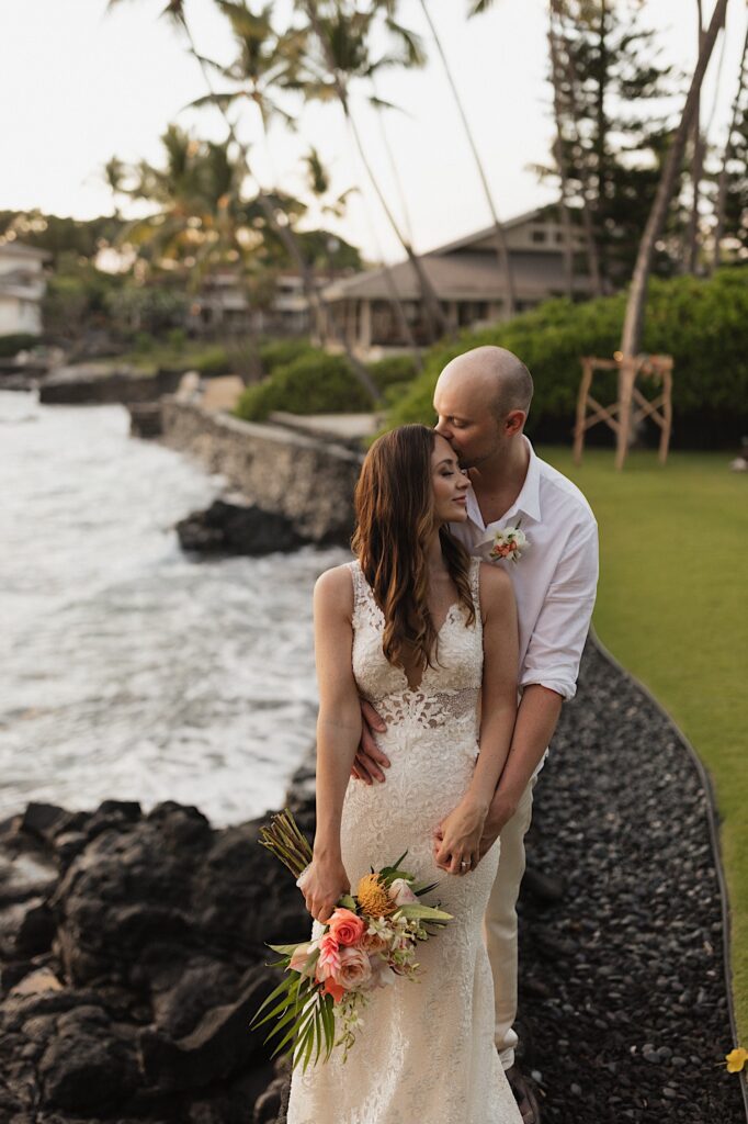 A bride looks over her shoulder as the groom behind her kisses her on the head while the two stand in a backyard next to the ocean