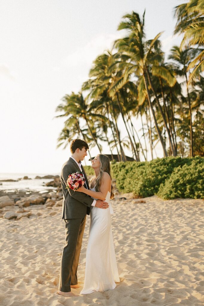A bride and groom on a beach face one another and smile while embracing during sunset with the ocean and palm trees behind them