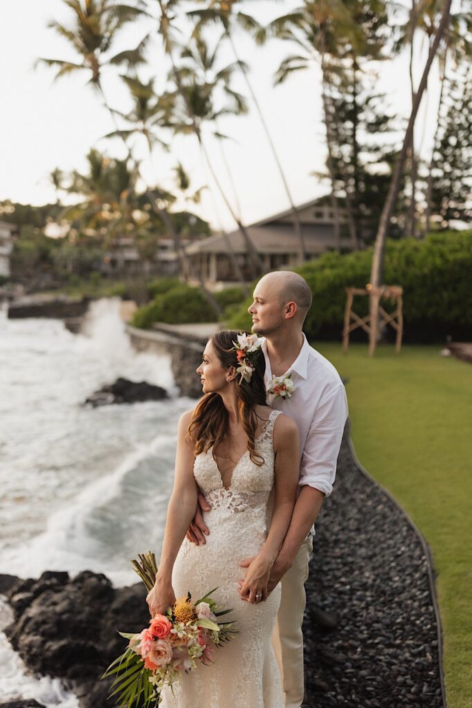 A bride stands with the groom standing behind her as the two look out at the ocean to the left while standing in a backyard