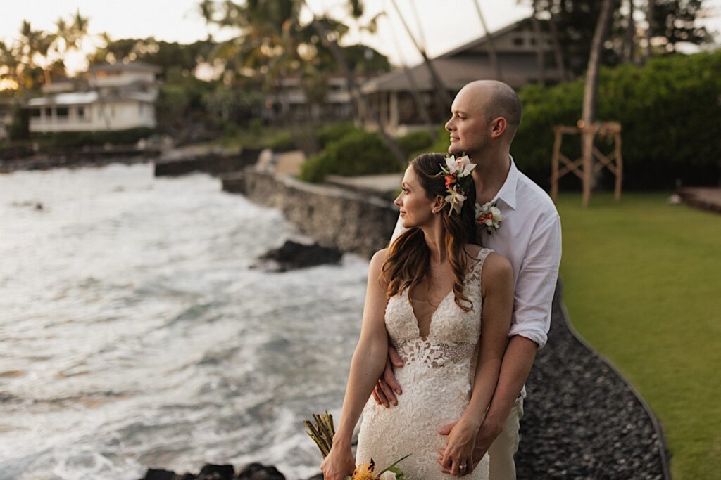 A bride stands with the groom behind her as they hold hands and look out at the ocean to the left after their intimate wedding ceremony on Hawaii's big island