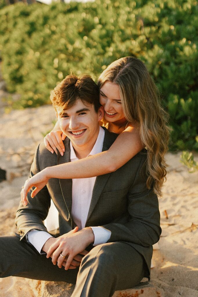 A groom sits on the beach while the bride crouches behind him and hugs him, both are smiling and the groom is looking at the camera, the sun is setting as well