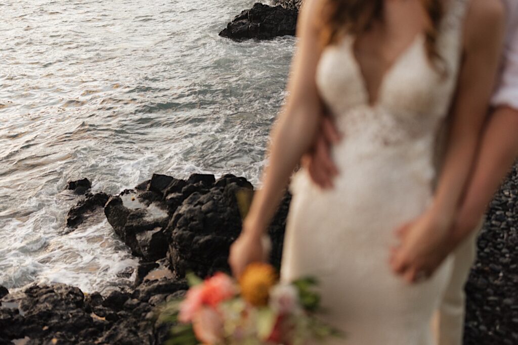Waves crash on the rocks below a bride and groom who are blurry in the foreground, the groom is standing behind the bride and holding her
