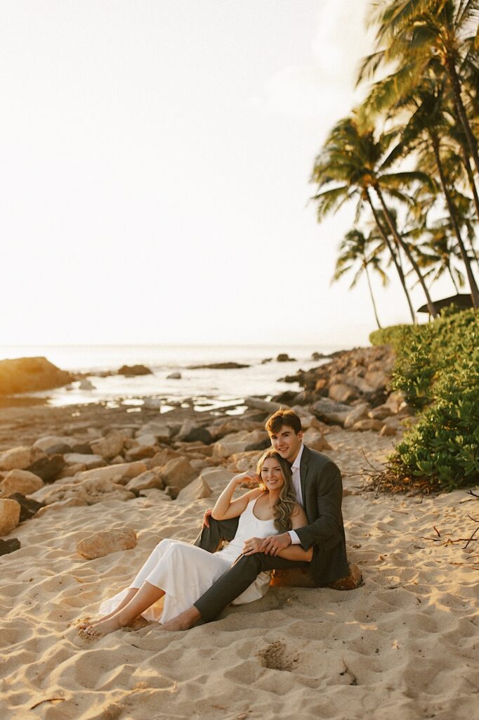 A bride sits on the beach between the groom's legs as they both smile at the camera while the sun sets with the ocean and palm trees behind them