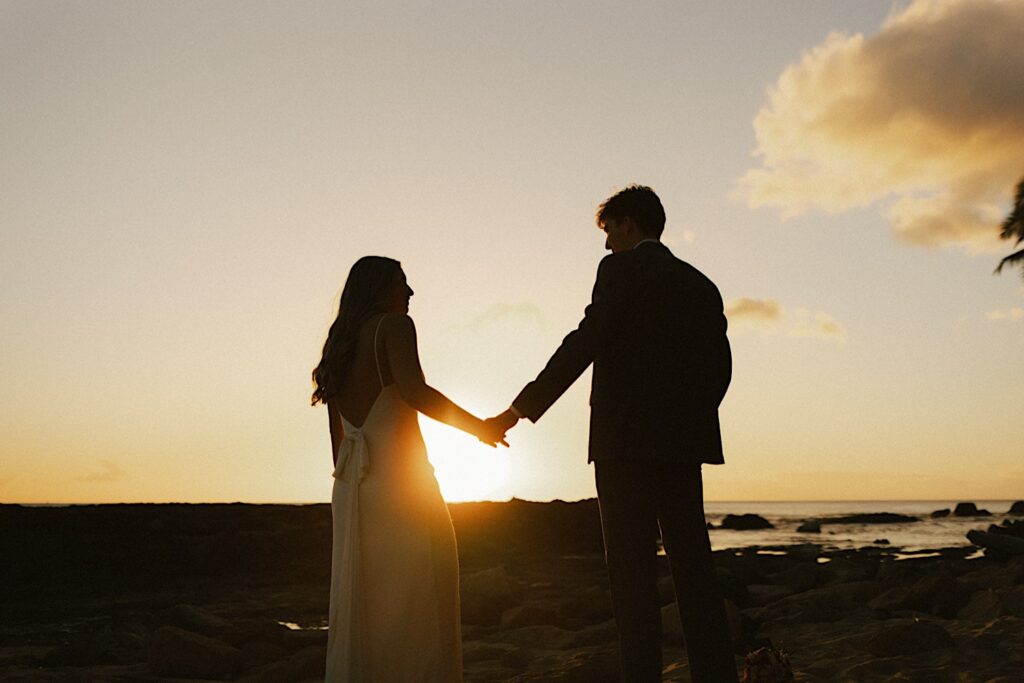 A bride and groom hold hands facing away from the camera while standing on a beach and looking at one another, in the background is the ocean and the sun setting on it during their elopement day at Lanikūhonua on Oahu