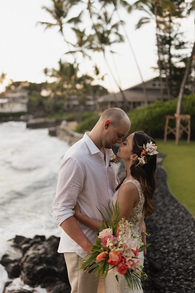 A bride and groom face one another and embrace while closing their eyes and touching noses together while standing in a backyard next to the ocean
