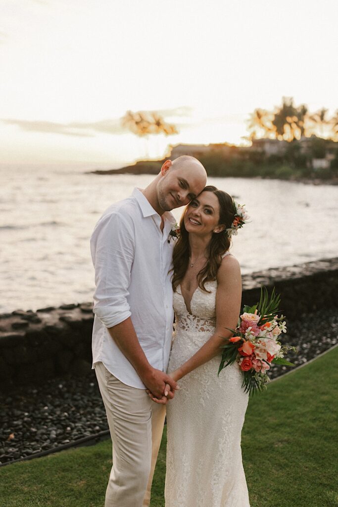 A bride and groom stand side by side and touch heads together while smiling at the camera, behind them is the ocean at sunset