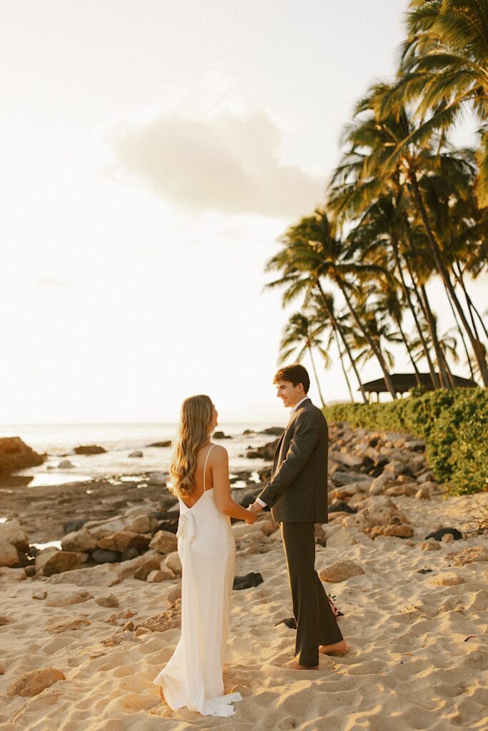 A bride and groom hold hands and look at one another while standing on a beach at sunset with the ocean and palm trees behind them