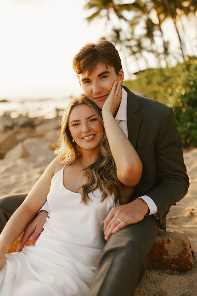 A bride sits on a beach between the groom's legs, both are smiling at the camera while the bride's hand rests on the groom's face as the sun sets with the ocean and palm trees behind them