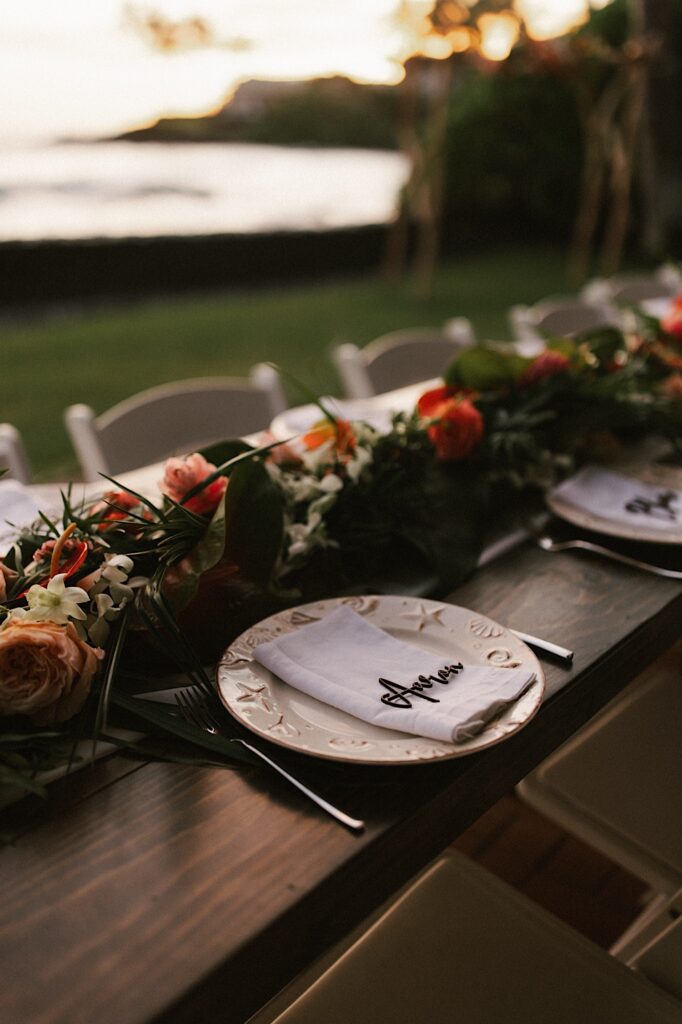 A napkin with a name on it sits on a table next to other plates with names set for a wedding reception, the table is decorated with flowers and leaves and the ocean is in the background
