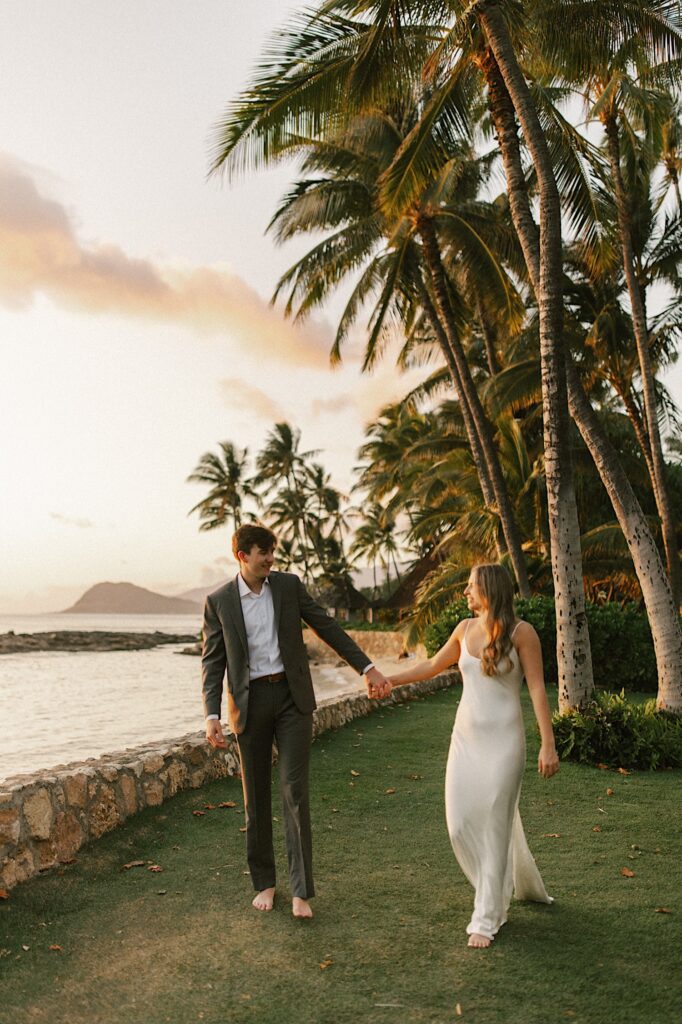 A bride and groom hold hands and smile at one another while walking towards the camera in the grass next to a beach, behind them are palm trees and the ocean as the sun sets