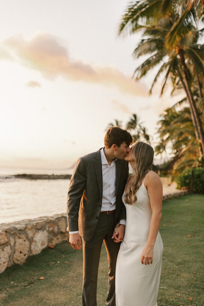 A bride and groom kiss one another while walking towards the camera in the grass next to a beach, behind them are palm trees and the ocean as the sun sets