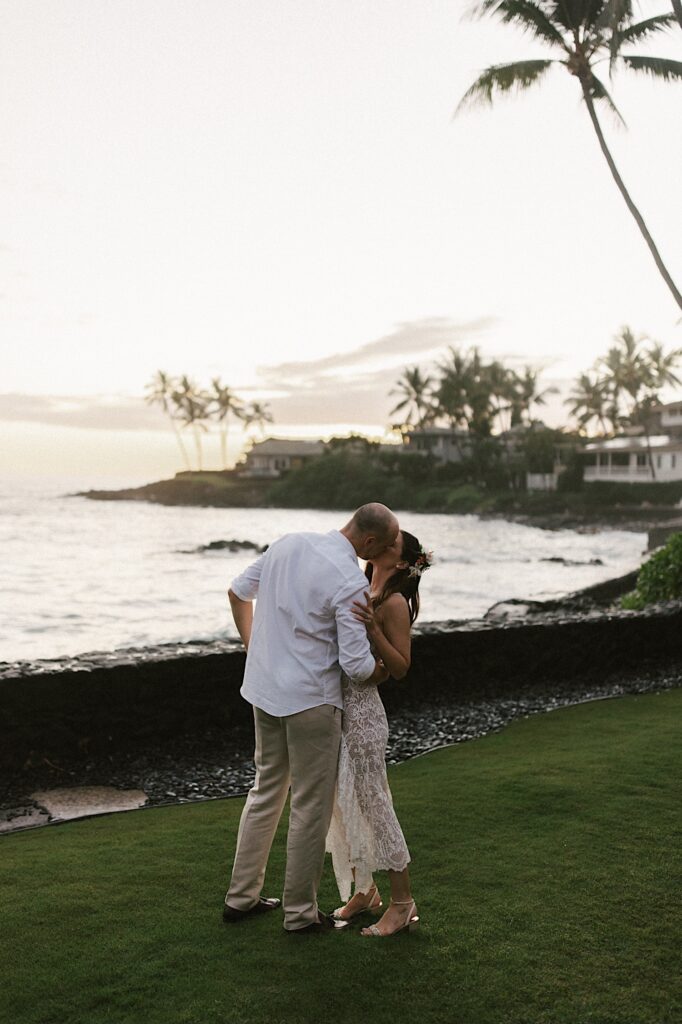A bride and groom kiss one another while standing in a backyard with the ocean and palm trees in the distance behind them