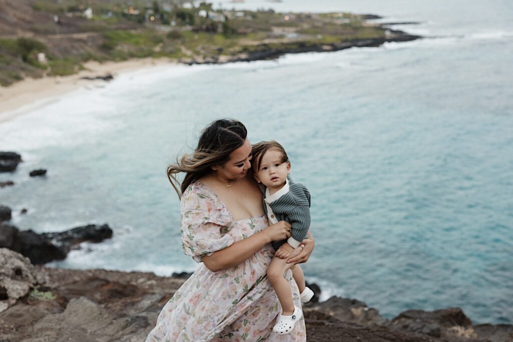 During a family session at Makapuu Lookout on Oahu a woman holds a young child and smiles at them while the child looks at the camera