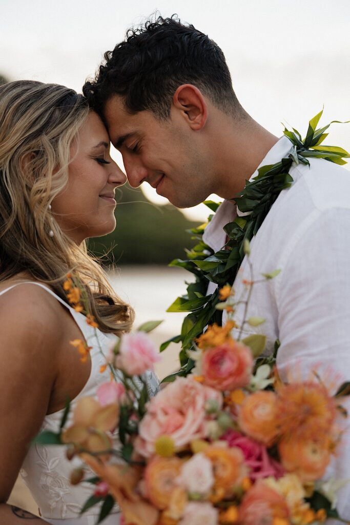 A bride and groom close their eyes and touch foreheads together while smiling, the groom is wearing a lei and the bride is holding a bouquet
