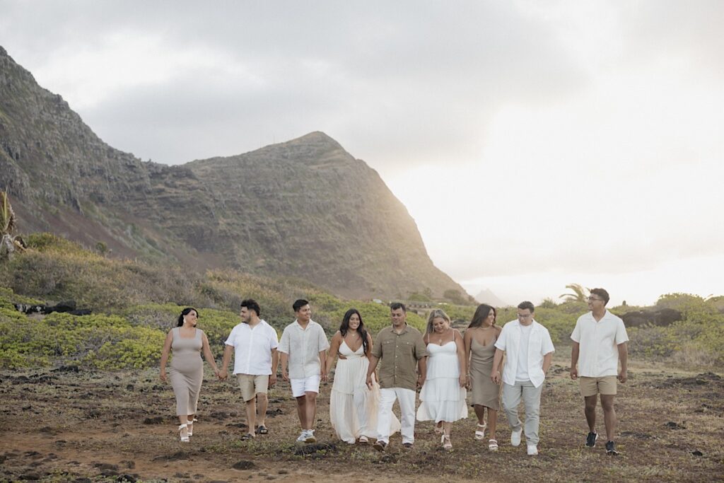 A family of 9 walk hand in hand towards the camera while in front of a mountain on Oahu
