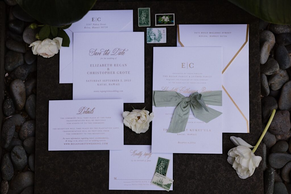A wedding day flatlay consisting of invites, flowers, and postcards sits on a pile of black stones