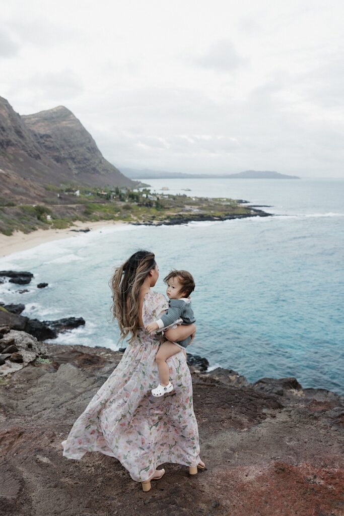 A woman holding a young child stands atop a cliff and looks out across the ocean and beach below