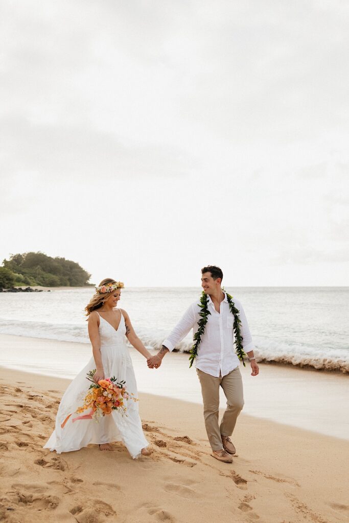 A bride and groom walk along a beach in Hawaii while smiling at one another, the groom is wearing a lei and the bride is holding a bouquet