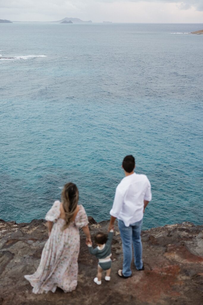 A man and woman stand with a small child standing in between them holding their hands, they're standing on a cliff looking out at the ocean below