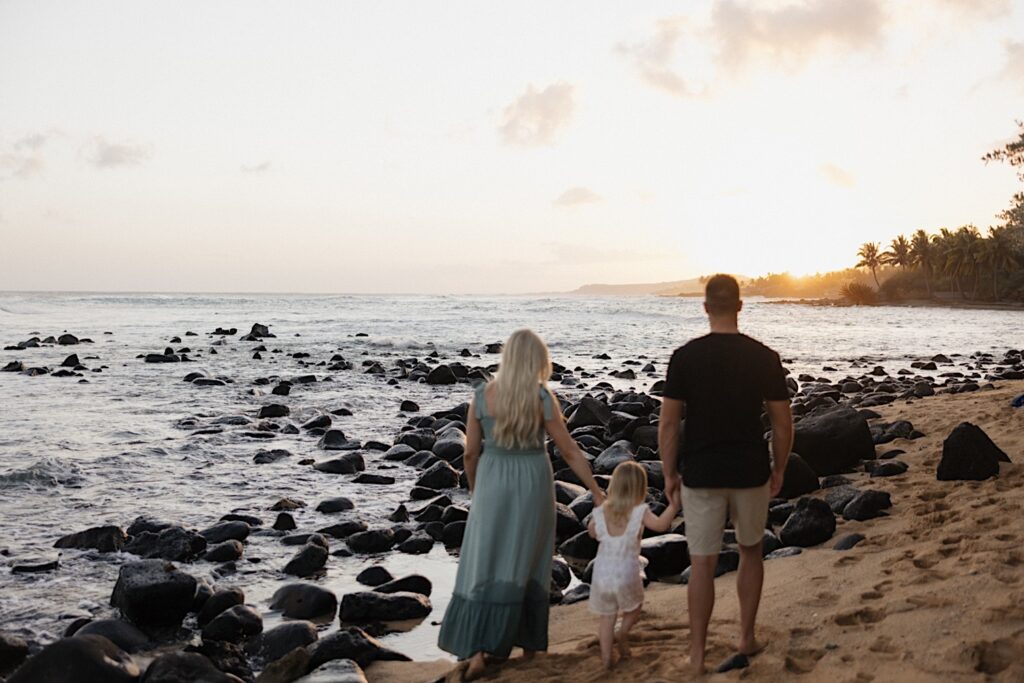 A family of 3 walk away from the camera towards the sunset while on a beach on Oahu during their family session