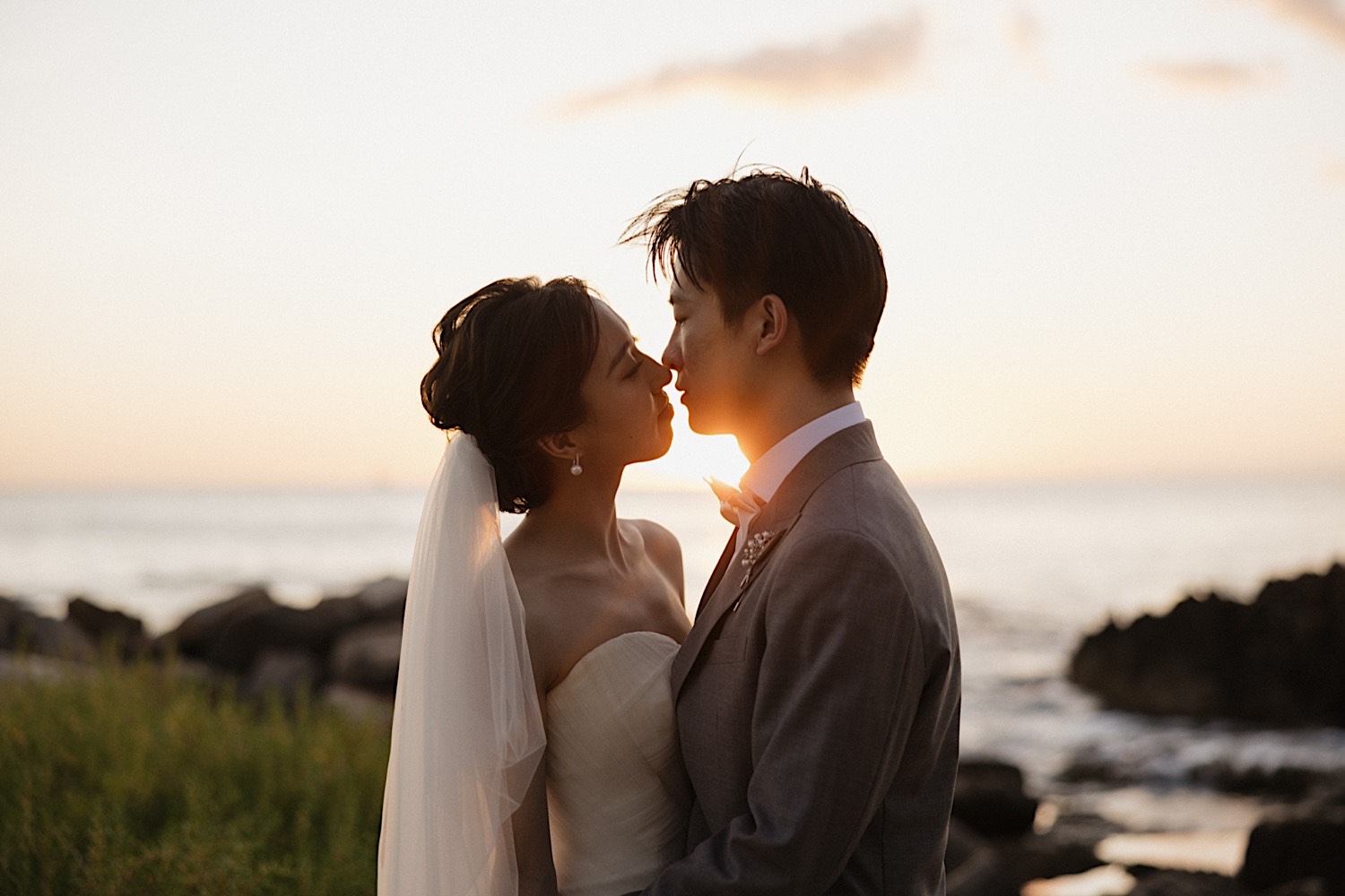 A bride and groom are about to kiss one another, behind them is the ocean and a sun setting directly between them during their elopement in Hawaii