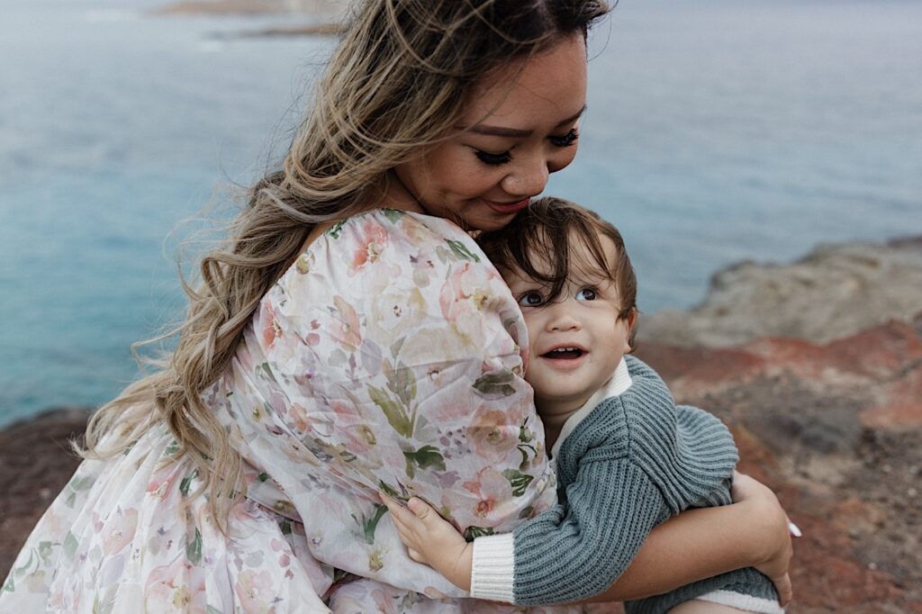 During a family session at Makapuu Lookout on Oahu a woman sits and smiles down towards her young child who is smiling up at something off camera