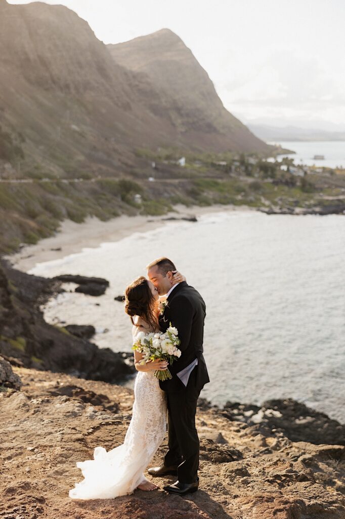 A bride and groom kiss one another while atop a cliff in Hawaii looking out over the ocean and mountains as the sun sets on them