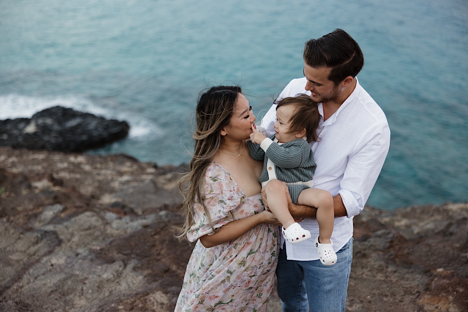 A mother and father hold their young child who is touching the mother's face, this is during their family session at Makapuu Lookout on Oahu