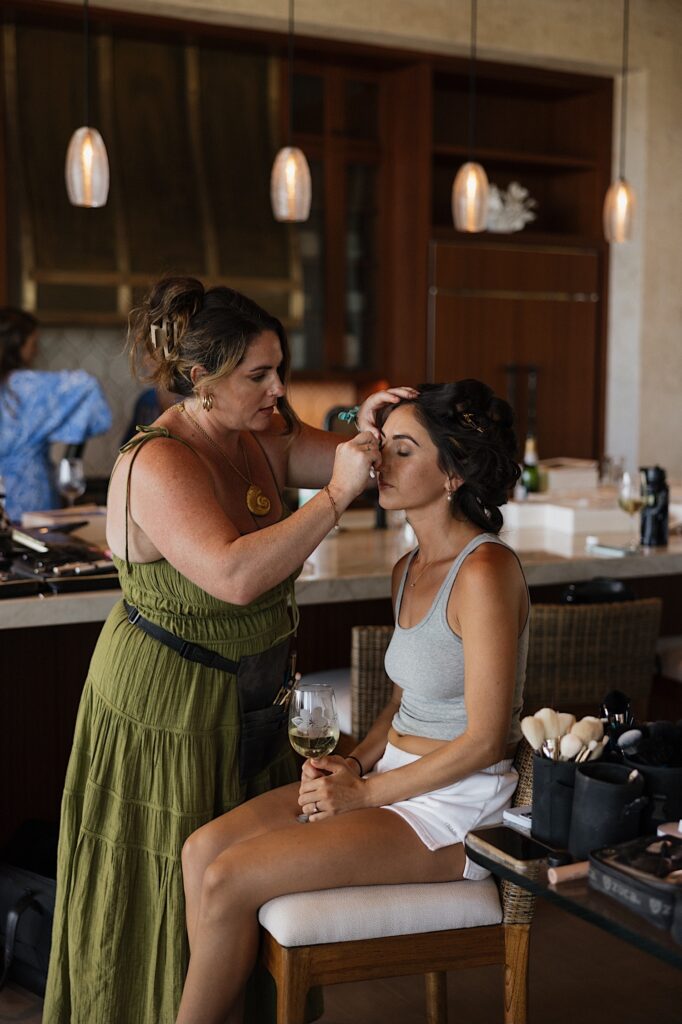 A bride sits in a chair with a glass of wine in hand as her hair and makeup artist works on her