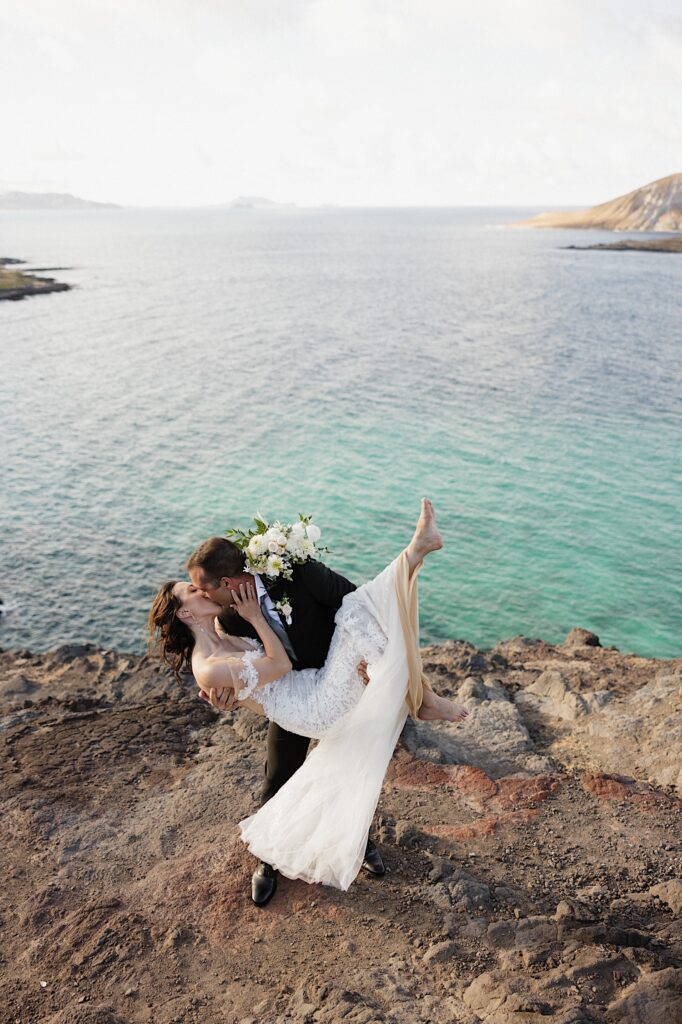 A bride is held by the groom who kisses her while the two stand atop a cliff looking out over the ocean in Hawaii