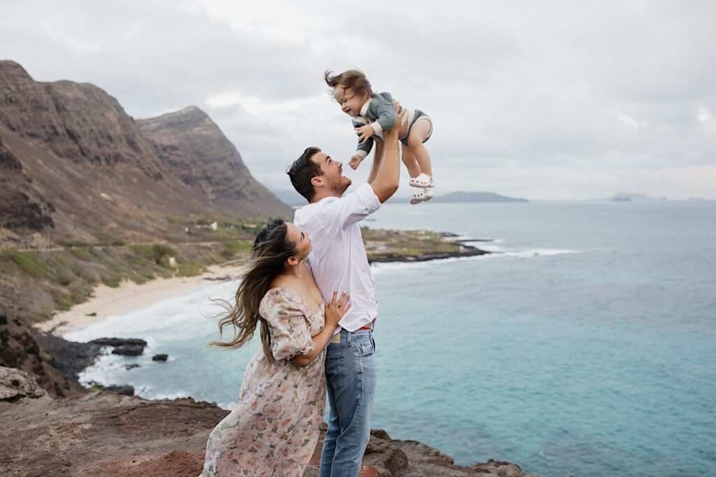 During a family session at Makapuu Lookout on Oahu a father lifts a child in the air as the mother watches from behind with the ocean and mountains of the island in the background