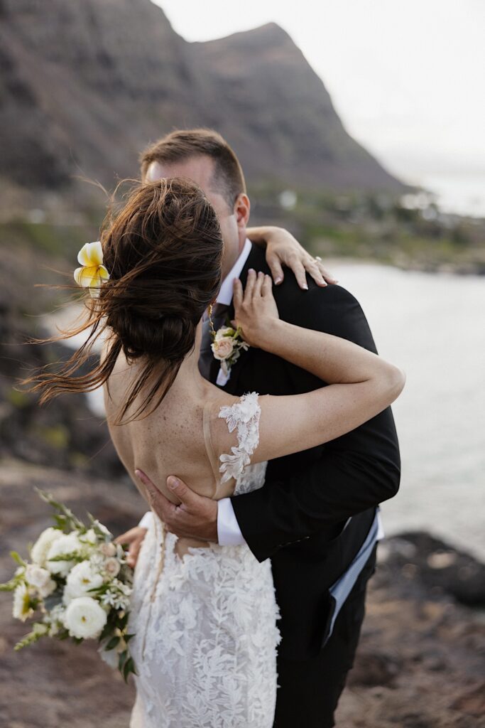 A bride with her back to the camera kisses the groom while the two stand atop a cliff in Hawaii that looks out over mountains and the ocean