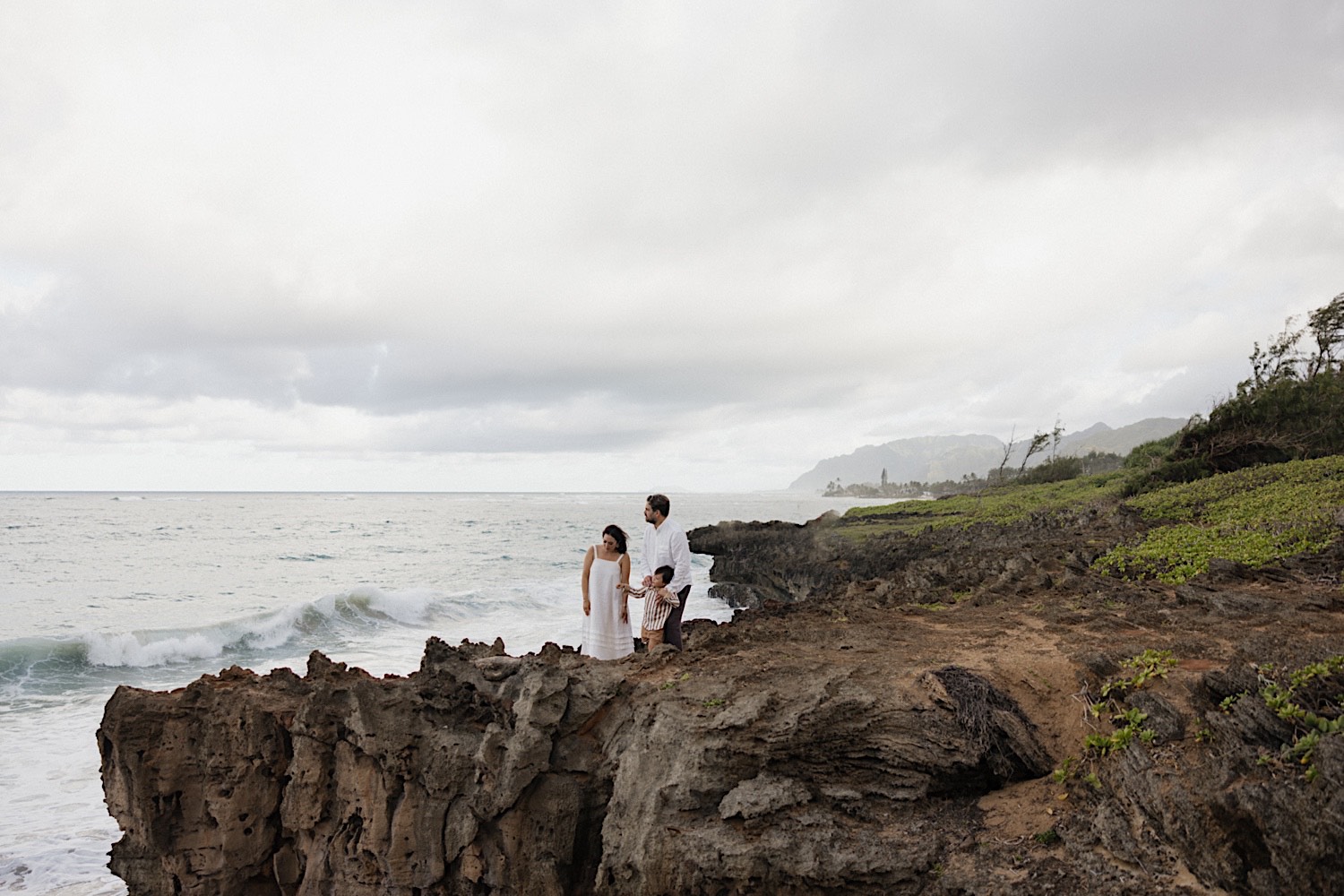 A family of 3 stand on a cliff next to the ocean during their family session on the island of Oahu