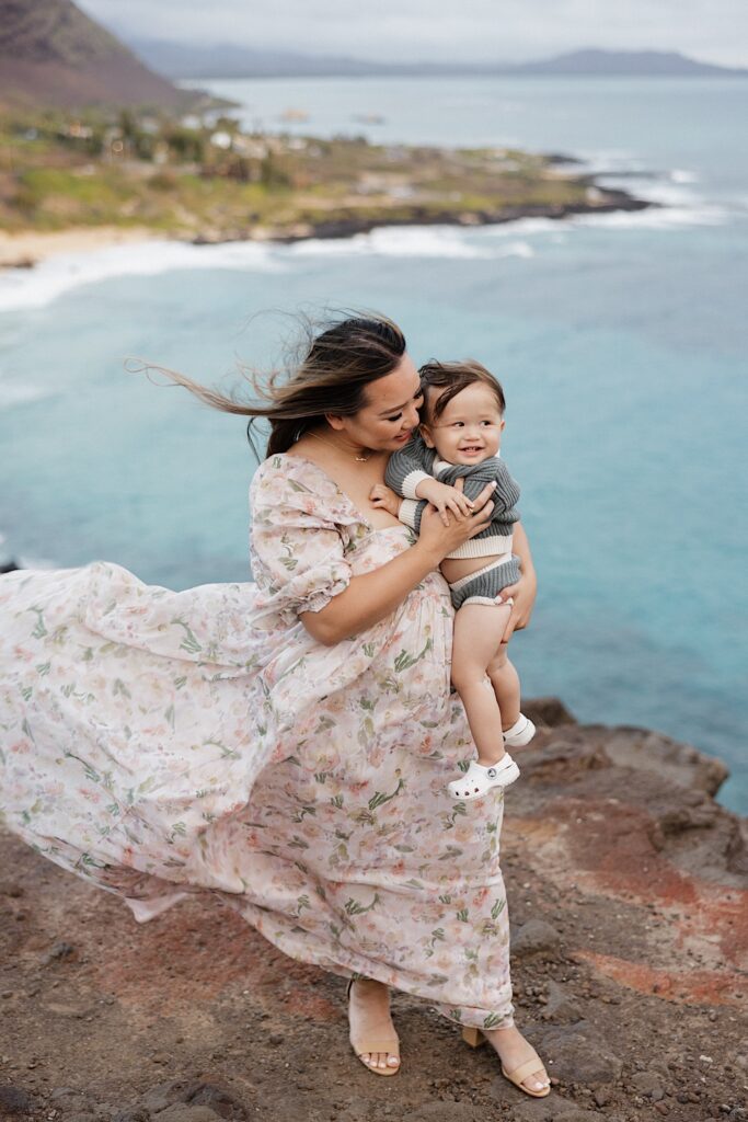 A mother holds her young child while standing atop a cliff with the ocean and island shore in the background
