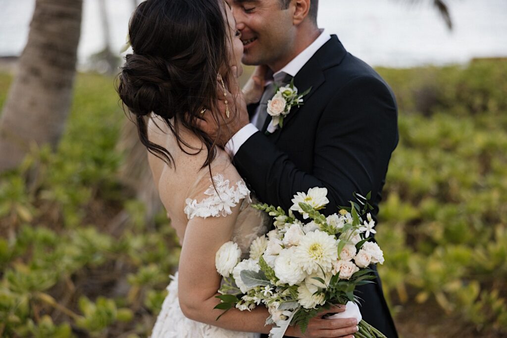 A bride and groom embrace to kiss one another during their elopement in Hawaii, the bride has her back to the camera and is holding a bouquet while the groom holds her face and smiles