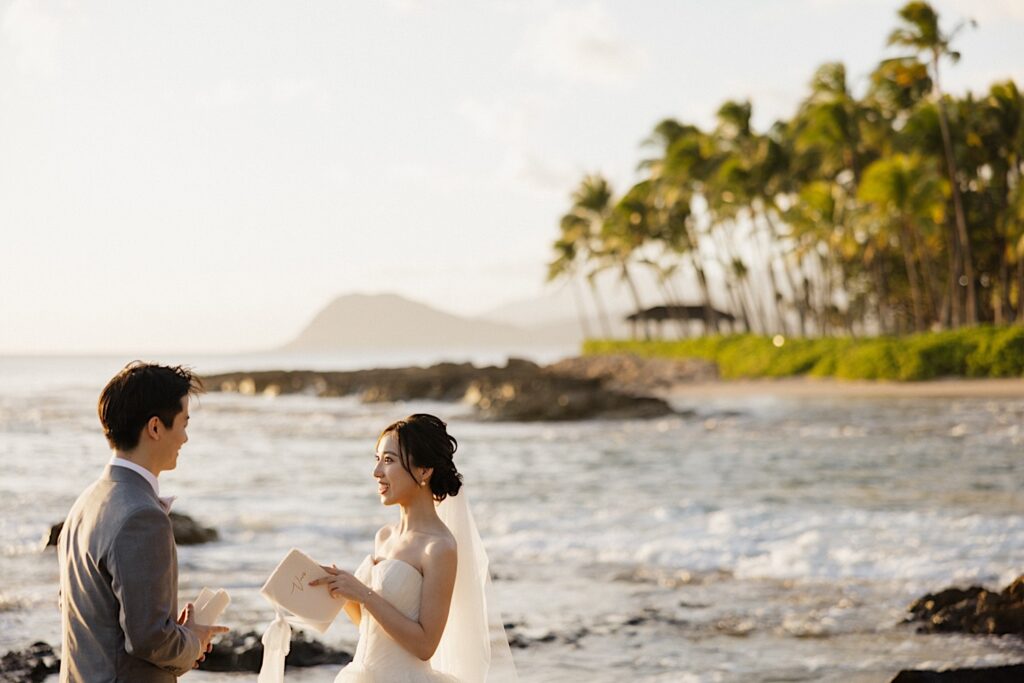 A bride and groom smile at one another while standing on a beach in Hawaii during their elopement with palm trees and the ocean in the background, each of them is holding their wedding vows