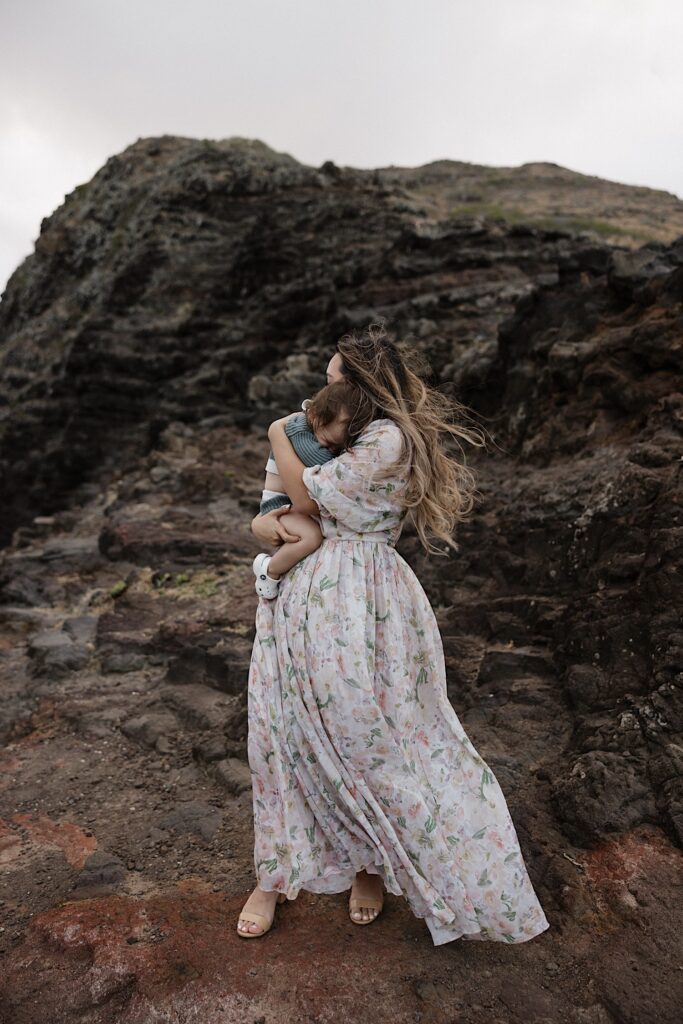 A woman holds and hugs her young child while standing on a rocky hill