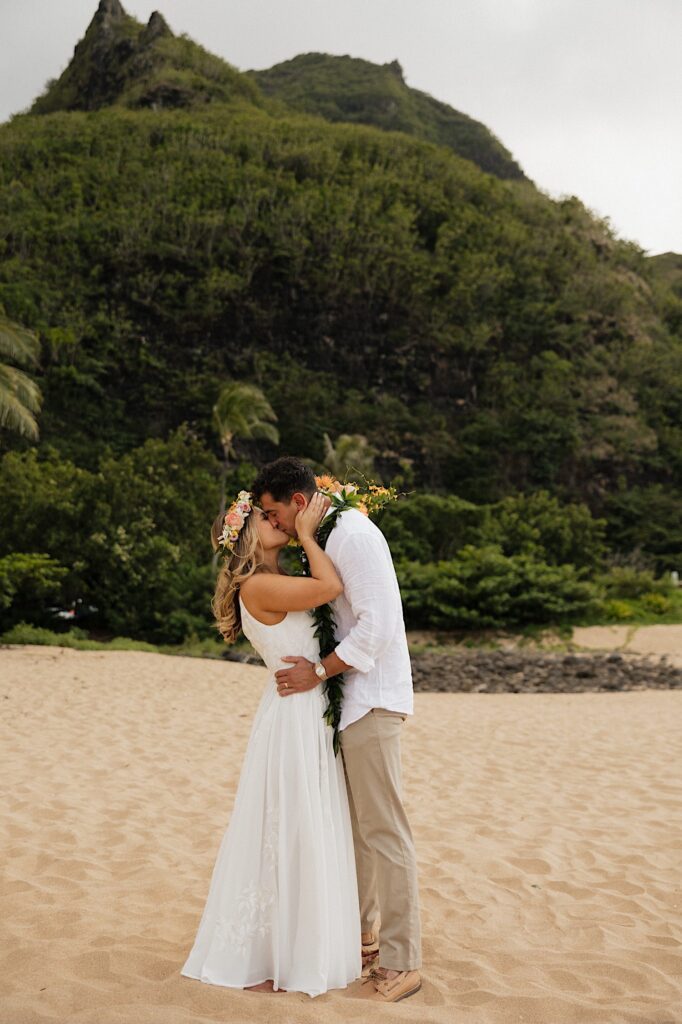 A bride and groom on a beach in Hawaii kiss one another in front of a large green mountain