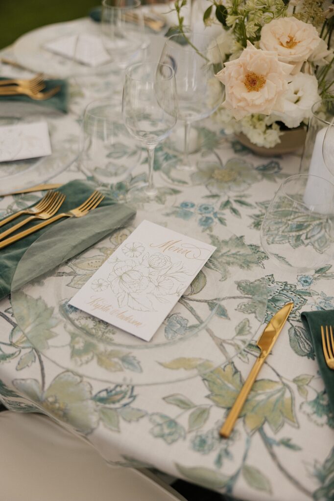A menu of a wedding sits on a clear plate on a table decorated for a wedding reception